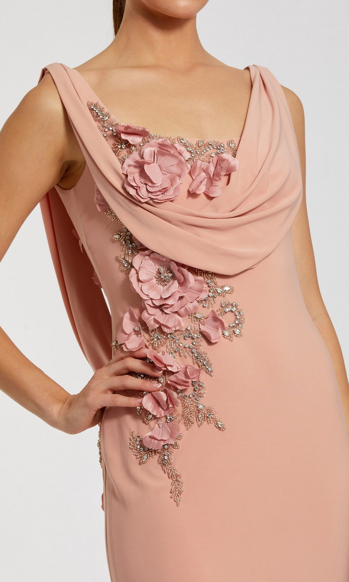 A woman wears the Mac Duggal 11910 Cowl-Neck Long Formal Dress, a sleeveless blush pink evening gown with a draped neckline and silver floral appliqué embellishments on the front.